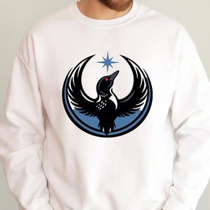May include: White sweatshirt featuring a black bird design with outstretched wings, a red eye, and a blue star. The bird is set within a blue and black circular frame. A casual, comfortable top.