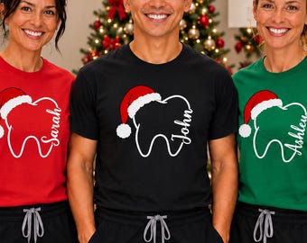 Könnte beinhalten: Drei kurzärmelige T-Shirts in Rot, Schwarz und Grün, jeweils mit einer weißen Grafik eines Zahns, der eine rote Weihnachtsmütze trägt. Die Shirts sind mit den Namen Sarah, John und Ashley in weißer Schreibschrift neben dem Zahn bedruckt.