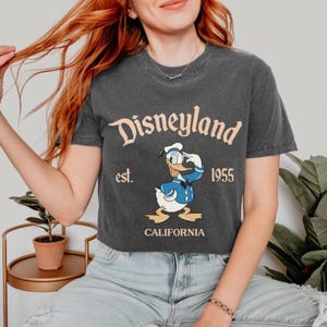 Pode incluir: Uma camiseta cinza escura cortada com um gr&aacute;fico vintage da Disneyland que apresenta o Pato Donald vestindo um traje de marinheiro e um chap&eacute;u. O texto diz "Disneyland est. 1955 California".