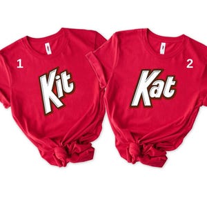 May include: Two red t-shirts with the words "Kit" and "Kat" in a white, brown, and black design. The shirts are knotted at the bottom. The shirts are short-sleeved.