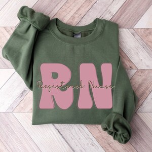 以下が含まれることがあります： ピンクの「RN」の文字と、金色の筆記体で「Registered Nurse」と書かれた緑色のスウェットシャツです。