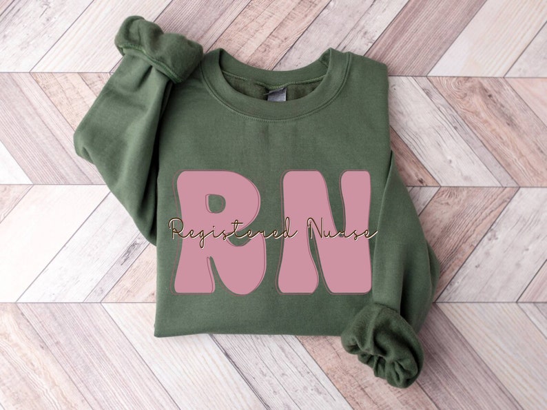 以下が含まれることがあります： ピンクの「RN」の文字と、金色の筆記体で「Registered Nurse」と書かれた緑色のスウェットシャツです。
