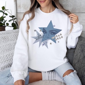 May include: White sweatshirt featuring a blue starburst design with the text "Silent Treatment" and "Freya Skye." The graphic includes a close-up of eyes. The sweatshirt is a casual, comfortable style.