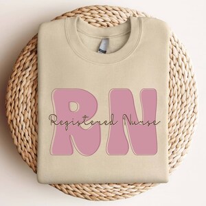 以下が含まれることがあります： ベージュのトレーナーに、ピンクの「RN」のグラフィックと、「Registered Nurse」というテキストが筆記体で書かれています。