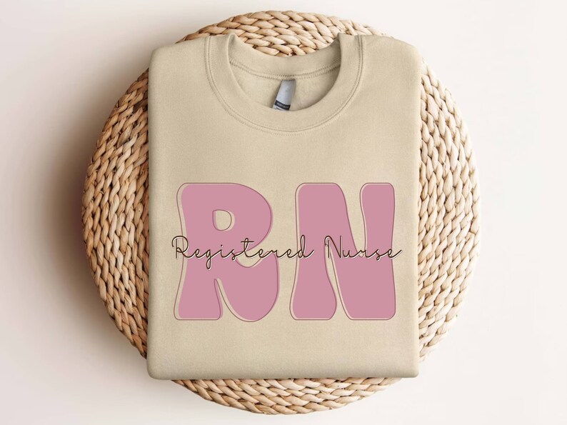以下が含まれることがあります： ベージュのトレーナーに、ピンクの「RN」のグラフィックと、「Registered Nurse」というテキストが筆記体で書かれています。