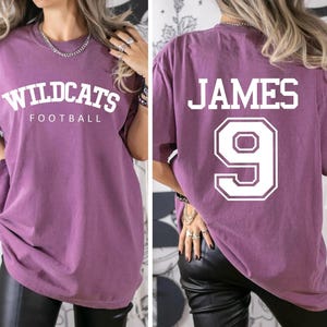May include: Purple oversized t-shirt with white text that says "Wildcats Football" on the front and "JAMES 9" on the back.
