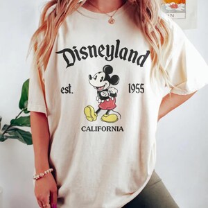 Pode incluir: Uma camiseta cor creme com um gr&aacute;fico preto e branco do Mickey Mouse. O texto "Disneyland est. 1955 California" est&aacute; impresso na camiseta.