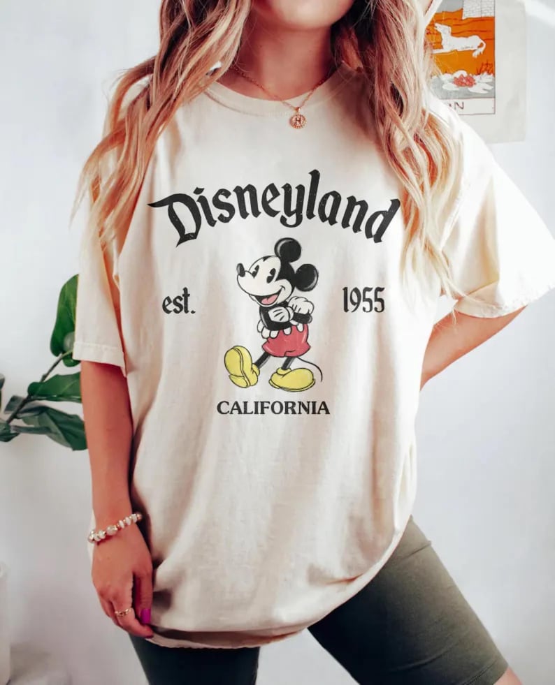 Pode incluir: Uma camiseta cor creme com um gr&aacute;fico preto e branco do Mickey Mouse. O texto "Disneyland est. 1955 California" est&aacute; impresso na camiseta.