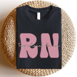 以下が含まれることがあります： 黒いTシャツにピンクの「RN」のデザインと、金色の筆記体で「Registered Nurse」の文字が書かれています。
