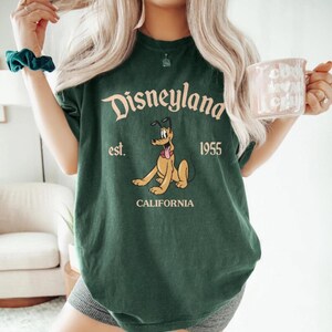 Pode incluir: Camiseta verde com uma imagem de desenho animado do Pluto, o c&atilde;o da Disney, e o texto "Disneyland est. 1955 California".