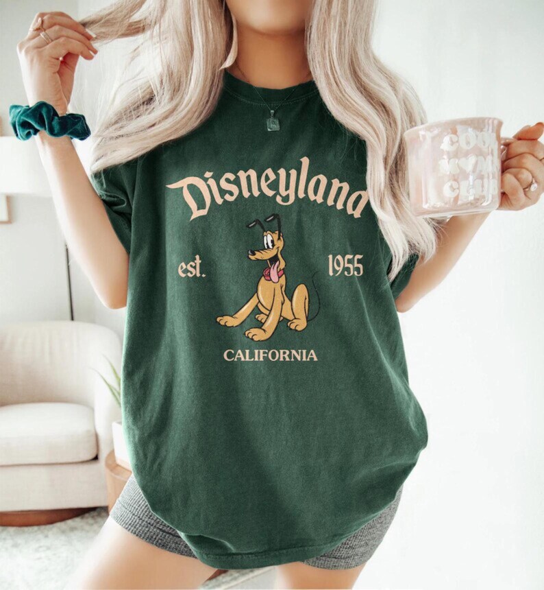 Pode incluir: Camiseta verde com uma imagem de desenho animado do Pluto, o c&atilde;o da Disney, e o texto "Disneyland est. 1955 California".