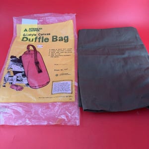 May include: An olive green canvas duffle bag folded flat next to its original clear plastic packaging. The packaging features the text American Camper G.I. style Canvas Duffle Bag, a graphic of the bag, and lists the size as 21 inches by 36 inches.