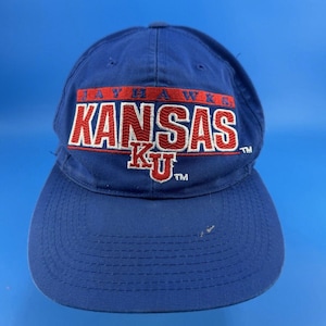 Puede incluir: Una gorra de béisbol azul real con texto bordado que dice JAYHAWKS en rojo sobre la palabra KANSAS en blanco con un borde rojo. Debajo, las letras KU están bordadas en rojo con un borde blanco, incluyendo los símbolos de marca registrada.
