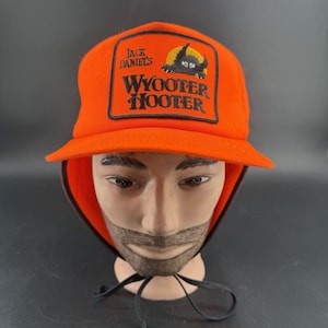 May include: A bright orange baseball cap with ear flaps and black chin straps. The front features a rectangular patch with the text Jack Daniel's and WYOOTER HOOTER in black, accompanied by an embroidered graphic of a black creature with yellow eyes.