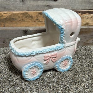 May include: A vintage ceramic baby carriage planter in pastel pink and white, accented with blue trim and a pink bow. The carriage features decorative wheels and a hood, perfect for small plants or decor.