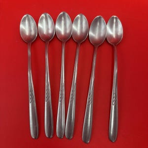 May include: Six stainless steel spoons with long handles and oval bowls are arranged on a red background. The handles have a decorative leaf design. The spoons are designed for stirring or serving.