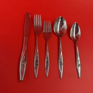 May include: A set of five silver-colored utensils on a red background. The set includes a knife, two forks, and two spoons. Each piece has a floral design on the handle.
