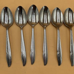 May include: A collection of eight stainless steel spoons displayed in a line. Each spoon features a teardrop-shaped bowl and a long, slender handle with a decorative element near the base. The spoons have a reflective, polished surface.