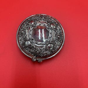 May include: A silver compact mirror with an ornate floral design. The mirror is closed and the design is visible on the front.