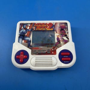 May include: A white handheld electronic game console featuring a Mega Man 2 graphic on the screen area. The device has a purple directional pad with a red cross, two red action buttons labelled Attack and Weapon, and a central speaker grille.
