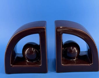 Può includere: Una coppia di fermalibri in ceramica marrone scuro, ciascuno caratterizzato da una cornice curva a forma di arco con una finitura liscia e lucida. Al centro dello spazio aperto di ogni arco si trova una sfera solida e compatta.