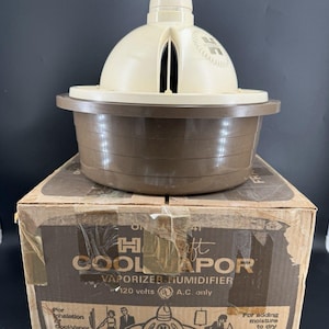 May include: A beige and brown vaporizer humidifier with a dome-shaped top and a one-gallon capacity. The device sits on its original cardboard box, which features the text "Hankscraft Cool Vapor Vaporizer-Humidifier 120 volts A.C. only".