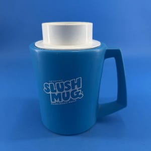 Puede incluir: Una taza para granizados de plástico azul brillante con borde superior y asa blancos. El frente presenta texto blanco que dice THE ORIGINAL SLUSH MUG FROZEN DESSERT MAKER. La taza tiene un cuerpo cilíndrico y un asa lateral resistente.