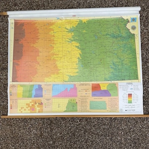 May include: A wall-mounted pull-down educational map of Kansas featuring a large central topographic map with color-coded elevation zones, surrounded by smaller inset maps detailing population density, land regions, and historical data.