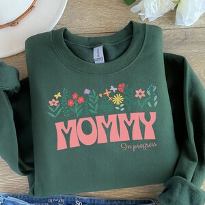 Puede incluir: Una sudadera verde oscuro con un gráfico rosa que dice "Mommy in progress" con un diseño floral sobre el texto.