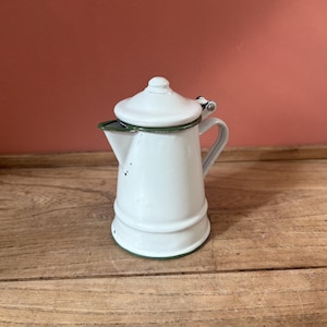 May include: A white enamel coffee pot with a tapered body, a hinged lid, and a side handle. The pot features a dark green rim along the lid, the spout, and the base. The surface has a smooth finish with minor dark spots near the spout area.