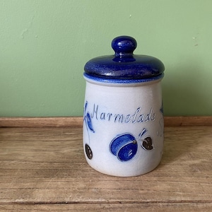 May include: A white ceramic jar with a blue lid and blue painted details. The jar is decorated with a blue and white design of plums and leaves. The word "Marmelade" is written on the front of the jar.