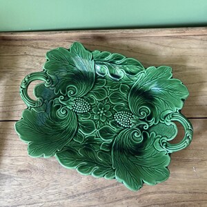 May include: A rectangular green ceramic serving tray featuring a detailed relief pattern of overlapping leaves, berries, and a central flower. The tray has two integrated handles on the sides and a glossy finish with darker green shading in the crevices.