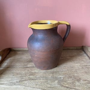 May include: A brown ceramic jug with a yellow rim. The jug is sitting on a wooden surface.
