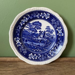 May include: A blue and white ceramic plate with a scalloped edge. The plate features a blue and white floral pattern and a central design of a bridge and trees.