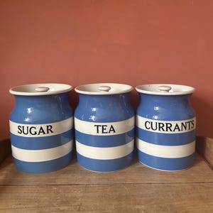 May include: Three blue and white striped ceramic kitchen storage jars with lids. Each jar is labelled with a black word: "SUGAR", "TEA", and "CURRANTS". The jars are arranged on a wooden surface.