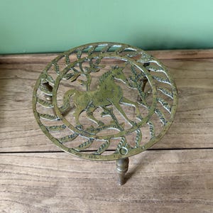 May include: A round brass trivet featuring an openwork design of a unicorn standing among stylised leafy branches. The circular metal piece is supported by three small, sturdy legs and displays a polished, metallic gold-toned finish.
