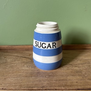 May include: A blue and white striped ceramic sugar jar. The word "SUGAR" is printed in black on the front. The jar has a white rim and is displayed on a wooden surface. A kitchenware item.