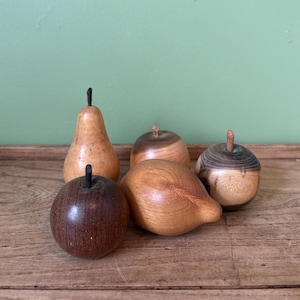 May include: A collection of four hand-carved wooden fruit sculptures, including a pear, a lemon, and two round apple-like shapes. Each piece displays distinct natural wood grain patterns, varying shades of brown, and small dark stems.