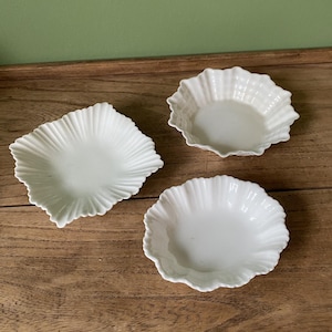 May include: Three white ceramic dishes with scalloped edges. The dishes are arranged on a wooden surface.