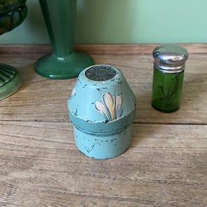 May include: A vintage, teal blue, round, decorative box with a floral design. The box has a silver lid with a floral design. The box is sitting on a wooden surface.
