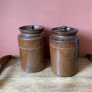 May include: Two antique brown stoneware jars with lids. The jars have a cylindrical shape with a decorative band around the middle. The lids have a knob on top. The jars are displayed on a wooden surface.