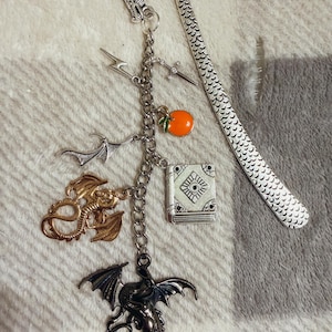 May include: A silver-toned metal bookmark featuring a dragon head hook and a textured scale pattern. A chain hangs from the hook, adorned with charms including a lightning bolt, a sword, an orange fruit, a bat, a book, and two dragon figures.