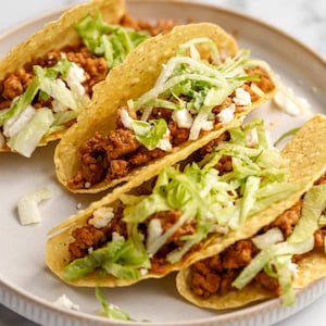 Può includere: Quattro tacos morbidi su un piatto bianco. Ogni taco è ripieno di carne macinata, lattuga tritata e formaggio sbriciolato.