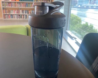 Può includere: Una bottiglia shaker nera con un corpo in plastica trasparente e un coperchio nero. La bottiglia è piena di un liquido scuro.