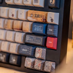 May include: Close-up of a black mechanical keyboard with custom keycaps. Keycaps feature text and graphics, including "Study in Scarlet," "221B Baker Street," and map designs. The keyboard has a variety of colors and textures.