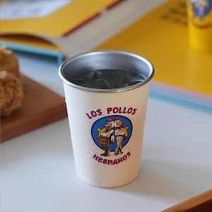 May include: A white metal cup with the text "LOS POLLOS HERMANOS" and a cartoon logo of two chickens. The cup has a silver rim and is filled with a dark liquid. The cup is on a white surface.
