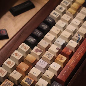 May include: A mechanical keyboard with a wooden frame and custom keycaps featuring various illustrations, symbols, and text. The keys are a mix of cream, brown, black, and textured finishes, with some displaying specific labels like Psychology.