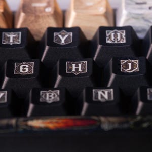 May include: Close-up of a black mechanical keyboard with custom keycaps featuring a medieval-style font and design. The keys are labelled with the letters G, H, J, B, and N.