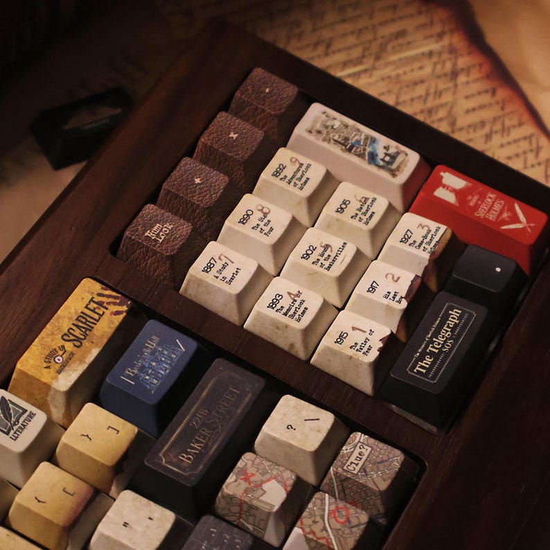 May include: A collection of custom keyboard keycaps with a Sherlock Holmes theme. The keycaps feature various designs, including book covers, maps, and text like "Baker Street" and "The Telegraph". The keycaps are arranged in a wooden tray.