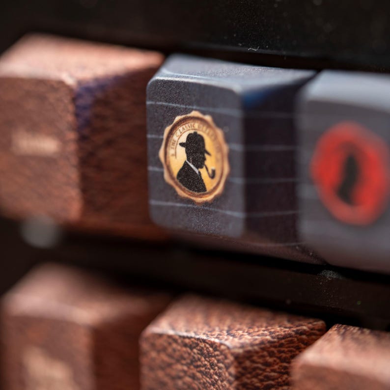 May include: A dark gray mechanical keyboard keycap featuring a circular gold emblem with a black silhouette of a person wearing a hat and smoking a pipe, surrounded by the text THE CLASSIC DETECTIVE, set against a background of textured brown keys.